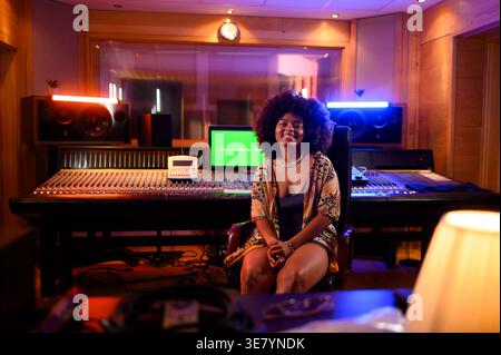 Lächelnde hispanische Musikerin mit Afro auf Stuhl vor Mischpult im Musikstudio Stockfoto