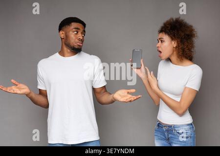 Schwarze Frau, die ihr Smartphone hält, bittet um eine Erklärung für ihren schwarzen Mann Partner vor grauem Hintergrund im Studio Stockfoto