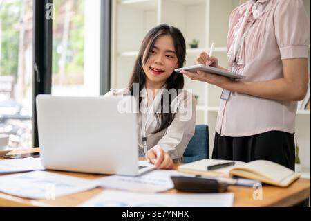 Asiatische Frau, die an einem Laptop arbeitet und eine Kollegin mit einem Tablet im Büro neben ihr steht Stockfoto