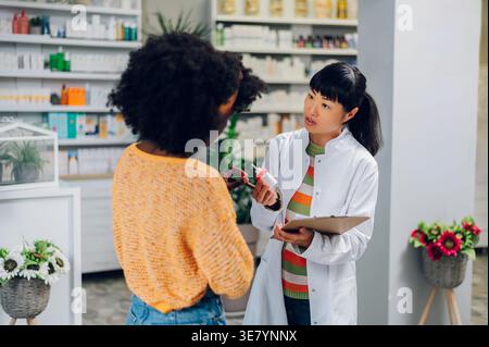Japanische Apothekerin mit Salbenspender und Klemmbrett, die mit schwarzer Patientin und Kundin in der Apotheke spricht Stockfoto