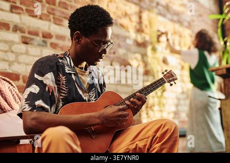 Schwarzer Mann mit Brille, der Akustikgitarre spielt, sitzt im Sessel gegen Ziegelwand im Wohnzimmer der Loft-Wohnung Stockfoto