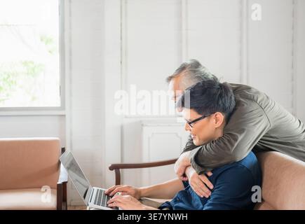 Asiatischer älterer Vater, der hinter ihm steht und einen Mann mittleren Alters umarmt, indem er zu Hause auf der Couch sitzt Stockfoto