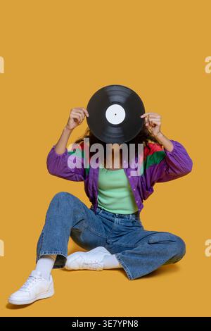 Afroamerikanerin, die das Gesicht mit Schallplatte bedeckt, sitzt drinnen auf dem Boden Stockfoto