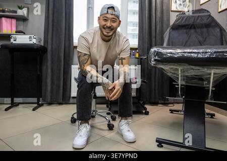 Lächelnder asiatischer Tätowierer, der tagsüber in seinem Tätowierstudio sitzt Stockfoto
