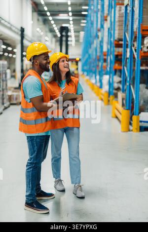 Schwarzer männlicher Arbeiter mit Schutzhelm und Sicherheitsweste, der Clipboard hält und Lagerbestand mit einer kaukasischen Kollegin verwaltet, während er im Lager steht Stockfoto