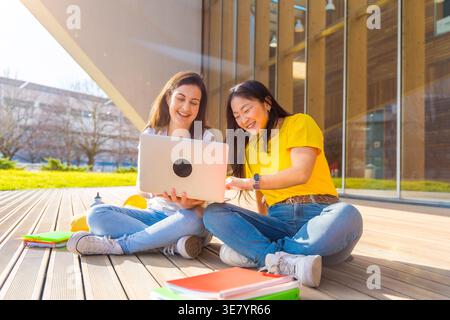 Fröhliche asiatische und kaukasische Studentin, die zusammen mit einem Laptop sitzt und mit überkreuzten Beinen auf einer Holzterrasse vor dem Universitätsgebäude auf der sonnigen... Stockfoto