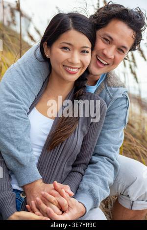 Lächelndes asiatisches Paar, das Hände hält und sich bei einem romantischen Date im Freien nahe beieinander im hohen Gras sitzt Stockfoto