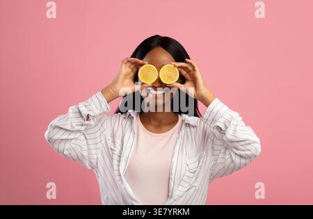 Schwarze Frau mit langen, geraden Haaren, die Augen mit Zitronenscheiben bedeckt, während sie im Studio vor rosa Hintergrund steht Stockfoto