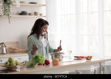 Schwarze Frau rührt Suppe im Topf, während sie an der Küchentheke mit Gemüse zu Hause steht Stockfoto