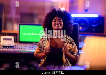 Fröhliche hispanische Musikerin mit Afro, die mit Händen auf ihrem Cardigan im Musikstudio sitzt Stockfoto