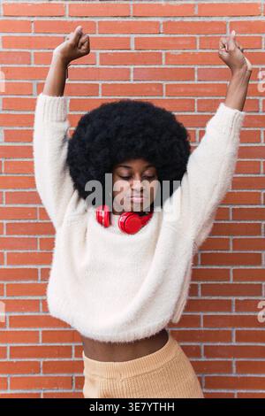 Schwarze Frau mit Afro-Kopfhörern am Hals, die Arme mit geschlossenen Augen ausstrecken und Spaß beim Tanzen gegen eine Ziegelwand haben Stockfoto