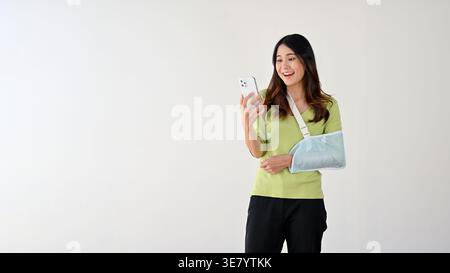 Lächelnde asiatische Frau mit Armschlinge mit Smartphone, das vor grauem Hintergrund steht Stockfoto