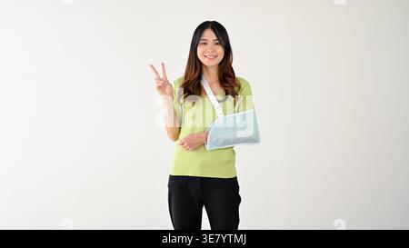 Lächelnde asiatische Frau mit Armschlinge mit Friedenszeichen, die vor grauem Hintergrund steht Stockfoto