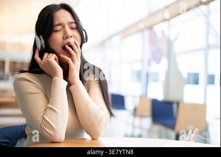 Verschlafene asiatische Frau mit geschlossenen Augen gähnt Musik mit Kopfhörern, die am Tisch im Café sitzen Stockfoto