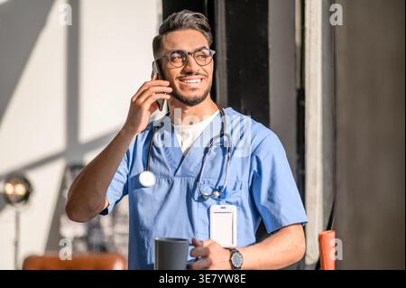 Lächelnder arabischer männlicher Arzt mit Stethoskop am Hals, Brille, Telefonanruf, Becher, der zu Hause wegblickt Stockfoto