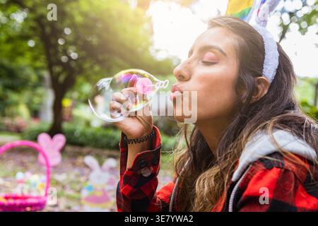 Hispanische Frau in Hasenohren Haarreif bläst Blasen mit geschlossenen Augen, während sie auf dem Boden im Park sitzt, um Ostern zu feiern Stockfoto
