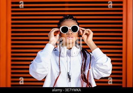 Schwarze Frau mit Kopfhörern am Hals, die Sonnenbrille an der orangefarbenen, gerippten Wand anbringen Stockfoto