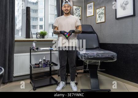 Lächelnder asiatischer Tätowierer mit Mütze und Latexhandschuhen, der verschiedene Tinten hält, bereitet sich auf die Tätowierung im Studio vor Stockfoto