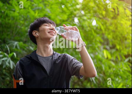 Lächelnder asiatischer Mann in Sportbekleidung, der Wasser in Flaschen trinkt, während er im Wald steht Stockfoto