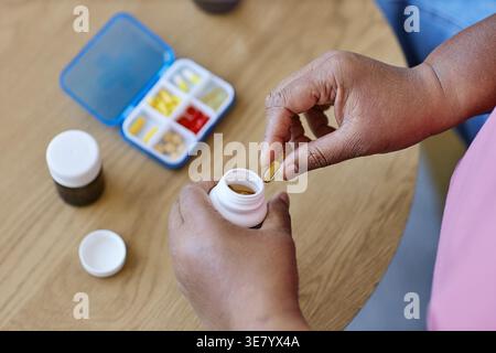 Hände einer schwarzen Frau mittleren Alters, die Kapseln auf Arzneimittelflasche nimmt, während sie zu Hause am Tisch im Wohnzimmer steht Stockfoto