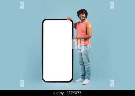 Bärtiger indischer Mann mit lockigem Haar, der geimpften Arm zeigt, während er in der Nähe eines großen Smartphones mit leerem Bildschirm vor blauem Hintergrund im Studio steht Stockfoto
