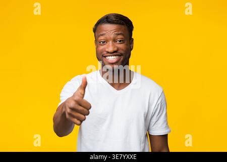Schwarzer Mann in weißem Hemd, der die Daumen hoch zeigt und vor gelbem Hintergrund im Studio steht Stockfoto