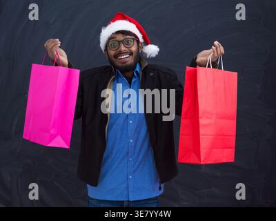 Fröhlicher indischer Mann mit Weihnachtsmann Hut mit Brille stehend, während er Einkaufstaschen vor dem Hintergrund der Tafel hält Stockfoto