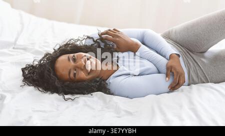 Lächelnde Schwarze Frau mit lockigem Haar, die sich umschließt und zu Hause im Bett liegt Stockfoto