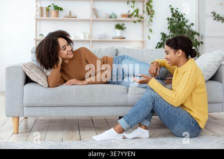 Traurige schwarze Frau, die auf dem Sofa liegt und mit einer Freundin spricht, während sie zu Hause im Wohnzimmer auf dem Boden sitzt Stockfoto