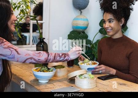 Hispanische und gemischte Freundinnen, die Spaß beim Essen und plaudern, während sie am Holztisch im Restaurant sitzen Stockfoto