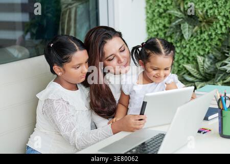 Indische Mutter und Töchter, die zusammen ein Tablet benutzen, sitzen am Tisch im Hinterhof Stockfoto