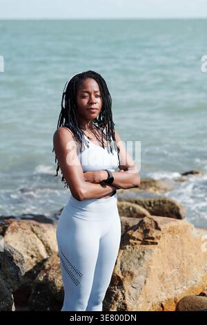 Schwarze Frau mit Dreadlocks im Fitness-Outfit mit überkreuzten Armen an sonnigen Tagen am Meer Stockfoto