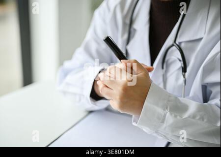 Asiatische Ärztin, die den Stift hält, während sie mit gefalteten Händen am Tisch in der Klinik sitzt Stockfoto
