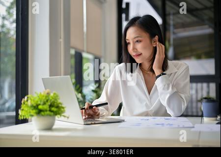 Asiatische Geschäftsfrau, die ihr Haar berührt, während sie am Laptop arbeitet und im Tischbüro sitzt Stockfoto