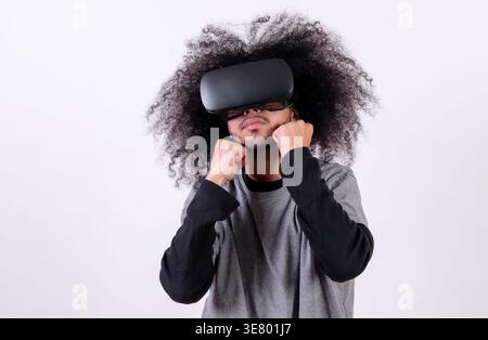 Hispanischer Mann mit lockigem Haar, der im Studio eine VR-Brille mit geballten Fäusten vor weißem Hintergrund trägt Stockfoto