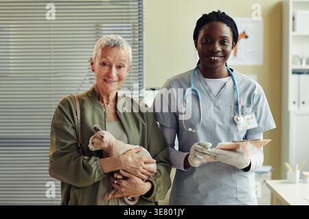 Lächelnde schwarze Tierärztin mit Klemmbrett neben der kaukasischen Seniorin, die tagsüber eine Katze in der Klinik trägt Stockfoto