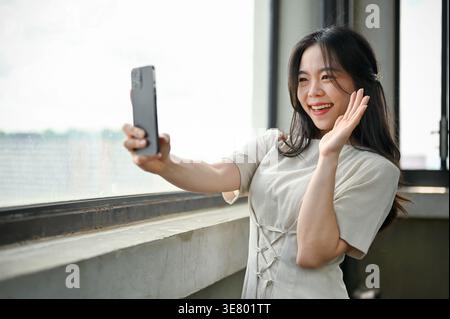 Eine attraktive Asiatin in einem schönen Kleid macht Selfies mit ihrem Smartphone, während sie in einem Gebäude am Fenster steht. Stockfoto