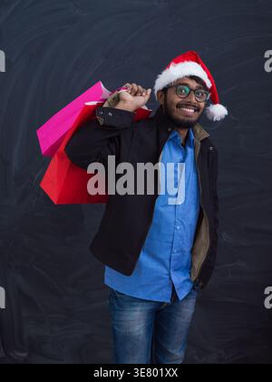 Fröhlicher indischer Mann mit Weihnachtsmann Hut mit Brille stehend, während er Einkaufstaschen vor dem Hintergrund der Tafel hält Stockfoto
