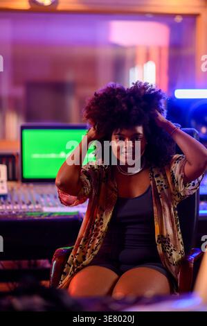 Lächelnde hispanische Musikerin, die ihr Afro-Haar anpasst, während sie im Musikstudio auf dem Stuhl sitzt Stockfoto