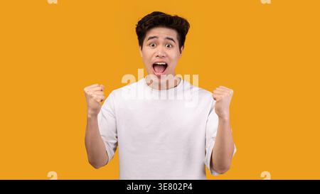Asiatischer Mann mit weißem Hemd, schreiend und geballt seine Fäuste, während er im Studio vor der orangen Wand steht Stockfoto