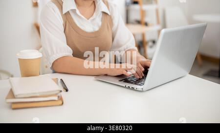 Zugeschnittenes Bild einer asiatischen Frau, die ferngesteuert mit einem Laptop arbeitet, der mit einer Kaffeetasse am Tisch sitzt und zu Hause Notizbücher verwendet Stockfoto