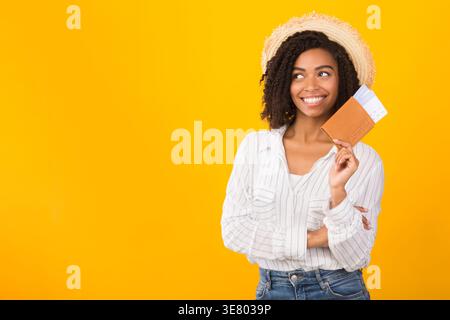 Schwarze Frau mit Strohhut mit Pass und Flugtickets, die vor gelbem Hintergrund im Studio stehen Stockfoto