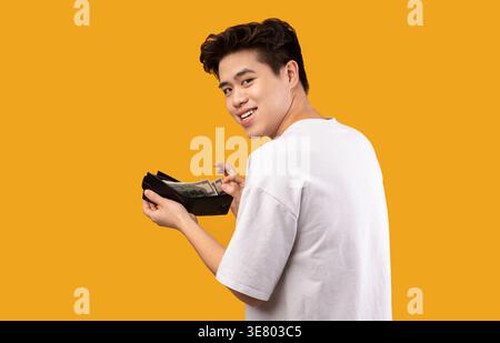 Asiatischer Mann mit weißem Hemd, der über die Schulter schaut und Geldbörse voller Geld hält, der vor der orangen Wand im Studio steht Stockfoto