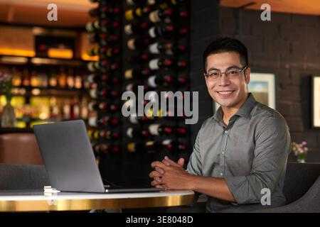 Asiatischer männlicher Unternehmer mit Brille am Tisch mit gefalteten Händen, der im Café am Laptop arbeitet Stockfoto