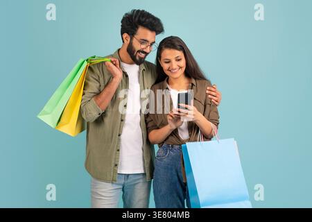 Lächelndes asiatisches Paar mit bunten Einkaufstaschen, das Smartphone benutzt, während es im Studio vor blauem Hintergrund steht Stockfoto