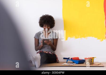 Schwarze Frau mit Afro, die auf dem Boden sitzt und sich entspannt, nachdem sie die Wand ihrer neuen Wohnung gemalt hat Stockfoto
