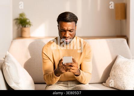 Seriöser Schwarzer Mann, der das Smartphone-Sofa im Wohnzimmer zu Hause benutzt Stockfoto
