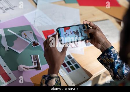 Hände eines Indianers, der ein Smartphone hält und Fotos von der Arbeit am Schreibtisch im Studio macht Stockfoto