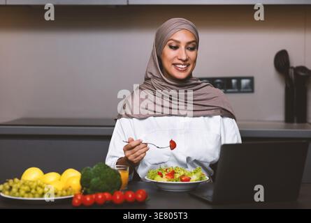 Lächelnde arabische muslimische Frau im Hijab, die auf den Laptop-Bildschirm blickt und Salat isst, während sie am Tisch sitzt mit Gemüse, einem Glas Orangensaft,... Stockfoto