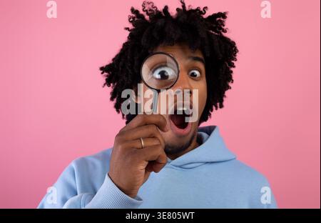 Schockierter Schwarzer Mann mit einem blauen Hoodie, der durch die Lupe blickt, während er im Studio gegen die pinkfarbene Wand steht Stockfoto
